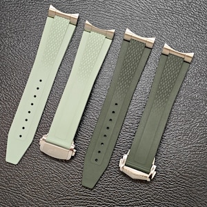 May include: Four watch straps in shades of green, displayed on a textured black surface. The straps feature a textured pattern and metal clasps. The straps range in colour from light green to a darker olive green.