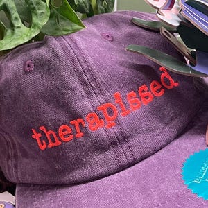 Könnte beinhalten: Eine lila Baseballkappe mit dem Wort "therapissed" in Rot gestickt. Die Kappe hat einen gebogenen Schirm und ein Etikett mit der Aufschrift "Embroidered Bear". Ein Aufkleber mit Text ist am Schirm befestigt.
