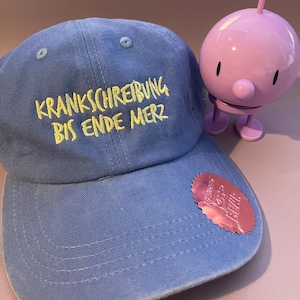 Könnte beinhalten: Eine blaue Baseballkappe mit gelbem Sticktext, der KRANKSCHREIBUNG BIS ENDE MERZ lautet. Die Kappe hat einen kleinen rosa, gezackten Aufkleber mit Text auf dem Schirm und steht neben einer kleinen, runden, hellvioletten Kunststofffigur.