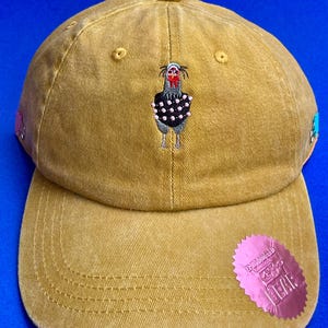 Pode incluir: Um boné de beisebol amarelo mostarda com um bordado de uma galinha vestindo uma peça preta com bolinhas. O boné inclui um adesivo circular rosa na aba com o texto EMBROIDERED BEAR e pequenos broches decorativos nas laterais.