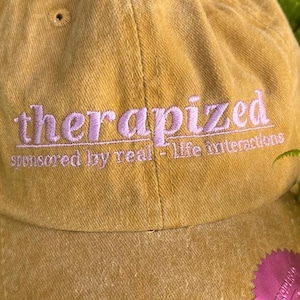 以下が含まれることがあります： マスタード色の野球帽。ピンク色で「therapized」の文字が刺繍されています。「sponsored by real-life interactions」のフレーズがメインテキストの下にあります。ピンク色のステッカーがツバに貼られています。