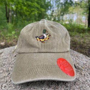 May include: Olive green baseball cap with an embroidered duck wearing a pizza-shaped floatie. The cap has a curved brim and a red patch on the side. The background is a blurred outdoor scene.