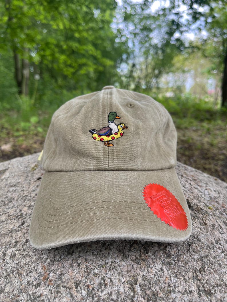 May include: Olive green baseball cap with an embroidered duck wearing a pizza-shaped floatie. The cap has a curved brim and a red patch on the side. The background is a blurred outdoor scene.