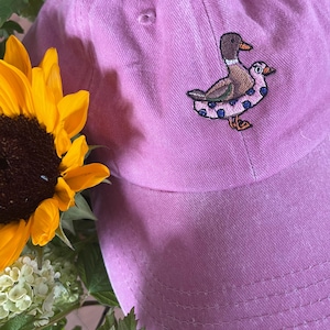 May include: A pink baseball cap with an embroidered design of two ducks. The ducks are brown, white, and blue. A yellow sunflower and white flowers are in the background.