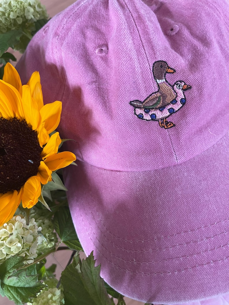 May include: A pink baseball cap with an embroidered design of two ducks. The ducks are brown, white, and blue. A yellow sunflower and white flowers are in the background.