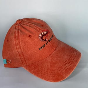May include: Orange baseball cap with a shrimp embroidered on the front. The text "KEEP IT SHRIMP" is embroidered below the shrimp. The cap has a curved brim and a teal-colored clip on the side.