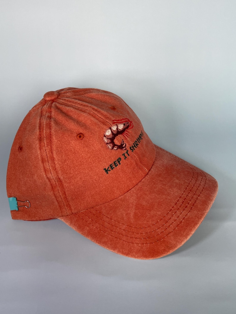 May include: Orange baseball cap with a shrimp embroidered on the front. The text "KEEP IT SHRIMP" is embroidered below the shrimp. The cap has a curved brim and a teal-colored clip on the side.