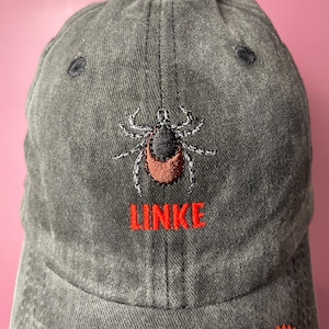 Könnte beinhalten: Eine anthrazitfarbene, verwaschene Baseballkappe mit einer gestickten Zecke in Schwarz, Weiß und Braun. Unter der Zecke ist das Wort LINKE in fetten, roten Großbuchstaben eingestickt. Die Kappe hat eine strukturierte Krone und Belüftungsösen.
