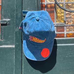 Könnte beinhalten: Blaue Baseballkappe mit dem Text "HOTmess" in einem flammenartigen Design. Die Kappe hat einen gebogenen Schirm und einen kleinen roten Aufnäher an der Seite. Die Kappe hängt an einer grünen Tür.