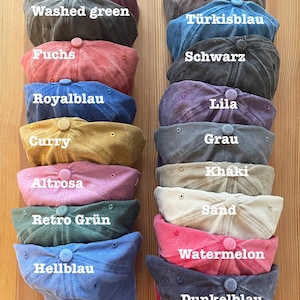 May include: A collection of baseball caps in various colors, including Bordeaux, Coffee, Washed Green, Fuchs, Royal Blue, Curry, Altrosa, Retro Green, Hellblau, and others. Each cap has a curved brim and a button on top. The caps are stacked on a wooden surface.