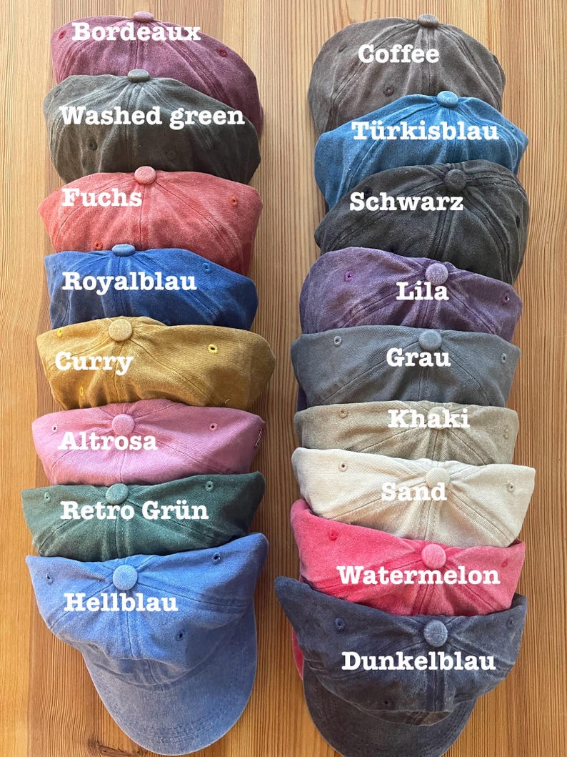 May include: A collection of baseball caps in various colors, including Bordeaux, Coffee, Washed Green, Fuchs, Royal Blue, Curry, Altrosa, Retro Green, Hellblau, and others. Each cap has a curved brim and a button on top. The caps are stacked on a wooden surface.