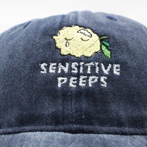 Könnte beinhalten: Marineblaue Baseballkappe mit dem gestickten Text "SENSITIVE PEEPS". Die Kappe hat ein gelbes Stickdesign einer weinenden Wolke mit einem grünen Blatt.