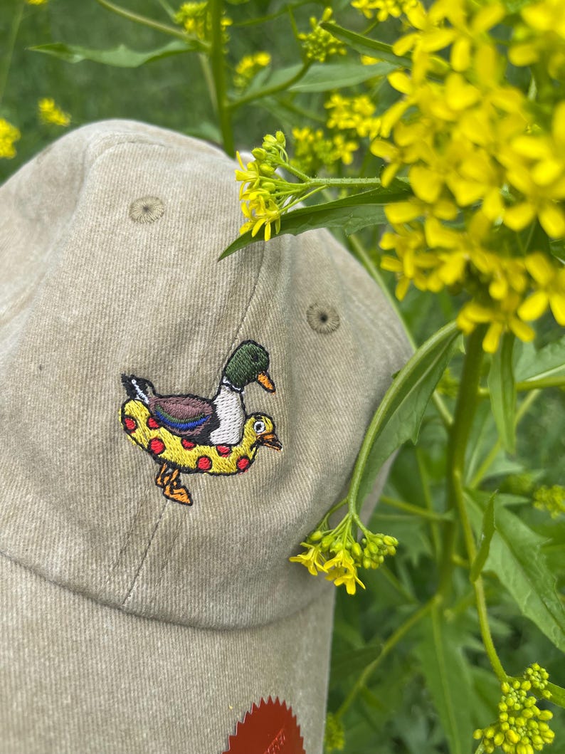 May include: A tan baseball cap with an embroidered design of a duck wearing a yellow and red inner tube. The cap is set against a backdrop of yellow flowers and green foliage.