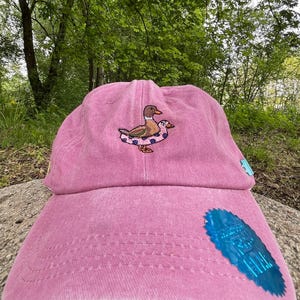 May include: A pink baseball cap with an embroidered design of a duck wearing a floatie. The cap has a curved brim and a blue tag on the side. The cap is sitting on a rock in front of a green background.