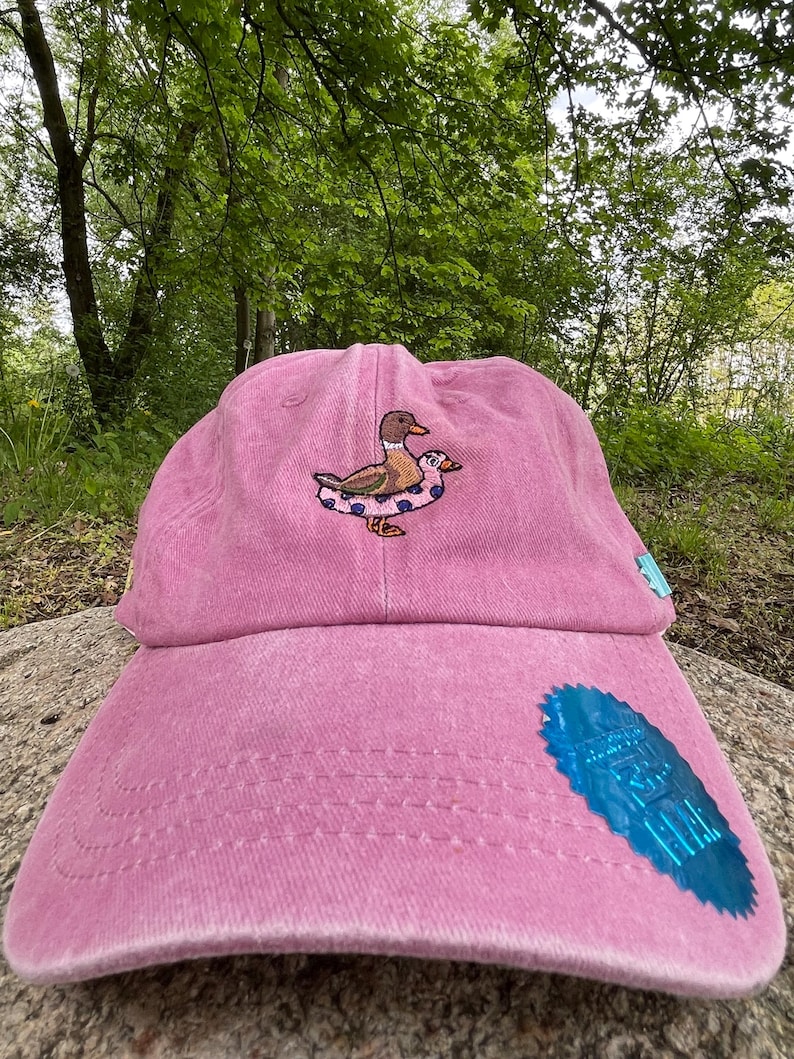 May include: A pink baseball cap with an embroidered design of a duck wearing a floatie. The cap has a curved brim and a blue tag on the side. The cap is sitting on a rock in front of a green background.