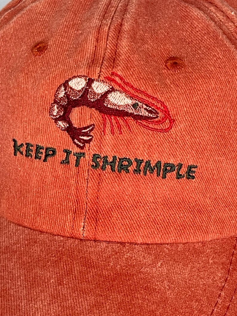 May include: Orange baseball cap with an embroidered shrimp design and the text "KEEP IT SHRIMPLE" in green. The cap has a curved brim and a distressed, vintage look.