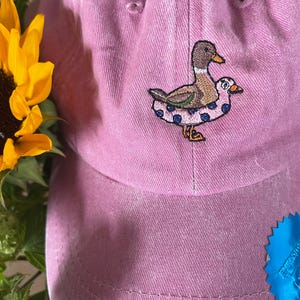 May include: A pink baseball cap with an embroidered duck design. The duck is brown, tan, and white, wearing a blue polka-dot inner tube. A yellow sunflower and green foliage are visible in the background.