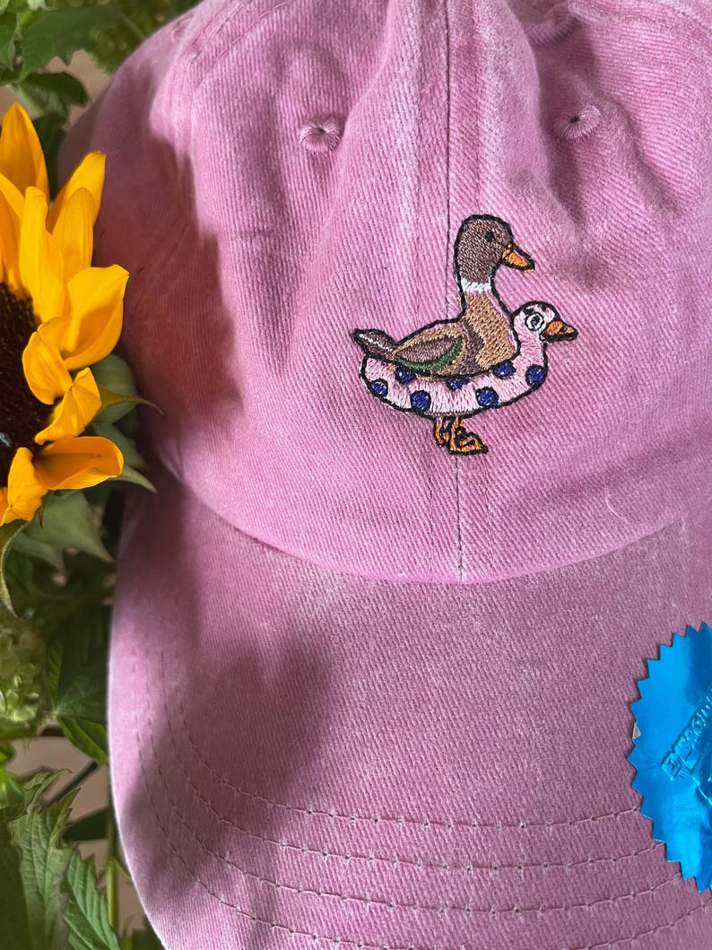 May include: A pink baseball cap with an embroidered duck design. The duck is brown, tan, and white, wearing a blue polka-dot inner tube. A yellow sunflower and green foliage are visible in the background.
