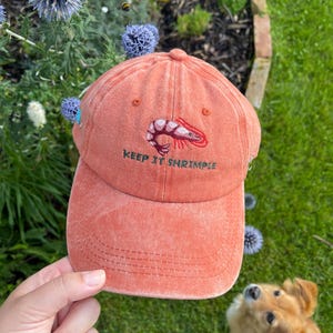May include: A coral-colored baseball cap with a curved brim. The cap features an embroidered shrimp graphic and the text "KEEP IT SHRIMPLE" in green. The hat is held in a hand, with a dog and flowers in the background.