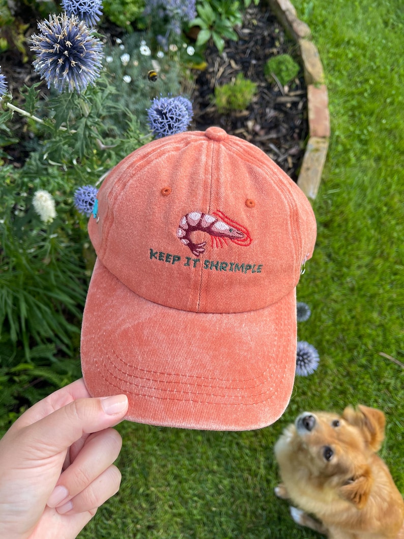 May include: A coral-colored baseball cap with a curved brim. The cap features an embroidered shrimp graphic and the text "KEEP IT SHRIMPLE" in green. The hat is held in a hand, with a dog and flowers in the background.