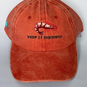 May include: Orange baseball cap with a shrimp graphic and the text "KEEP IT SHRIMPLE". The cap has a curved brim and a fabric strap closure. The cap is made of a soft, comfortable material.