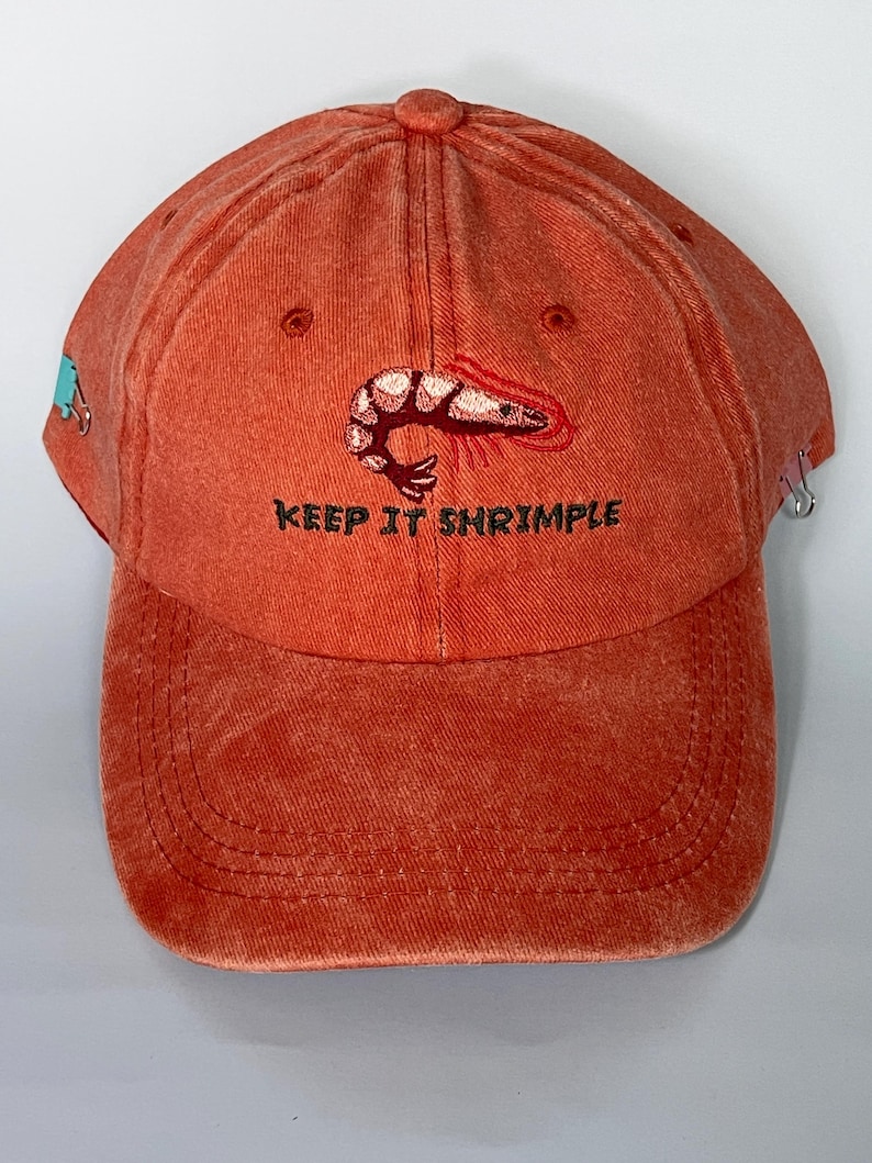 May include: Orange baseball cap with a shrimp graphic and the text "KEEP IT SHRIMPLE". The cap has a curved brim and a fabric strap closure. The cap is made of a soft, comfortable material.