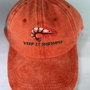 May include: An orange baseball cap with the embroidered phrase "KEEP IT SHRIMPLE" and a shrimp graphic. The cap has a curved brim and a button on top. The cap is made of a textured fabric.