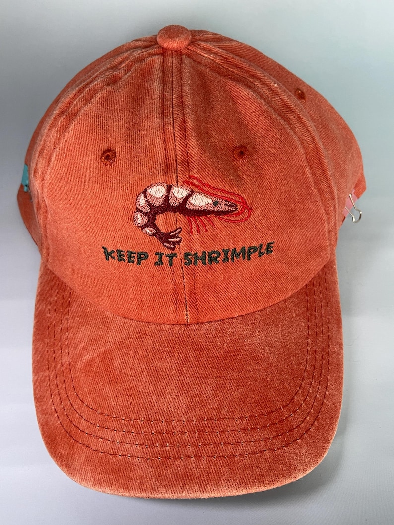 May include: An orange baseball cap with the embroidered phrase "KEEP IT SHRIMPLE" and a shrimp graphic. The cap has a curved brim and a button on top. The cap is made of a textured fabric.