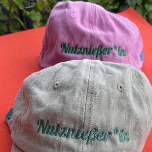 Könnte beinhalten: Zwei Baseballkappen, eine in Hellrosa und eine in Hellgrau, beide mit dem aufgestickten grünen Schriftzug "Nutznießer*in" auf der Vorderseite. Jede Kappe verfügt über gestickte Ösen an der Krone und eine strukturierte Stoffkonstruktion mit Schirm.