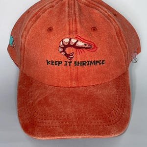May include: A burnt orange baseball cap with a curved brim. The cap features an embroidered shrimp design and the phrase "KEEP IT SHRIMPLE" in black lettering. A small blue design is on the side.
