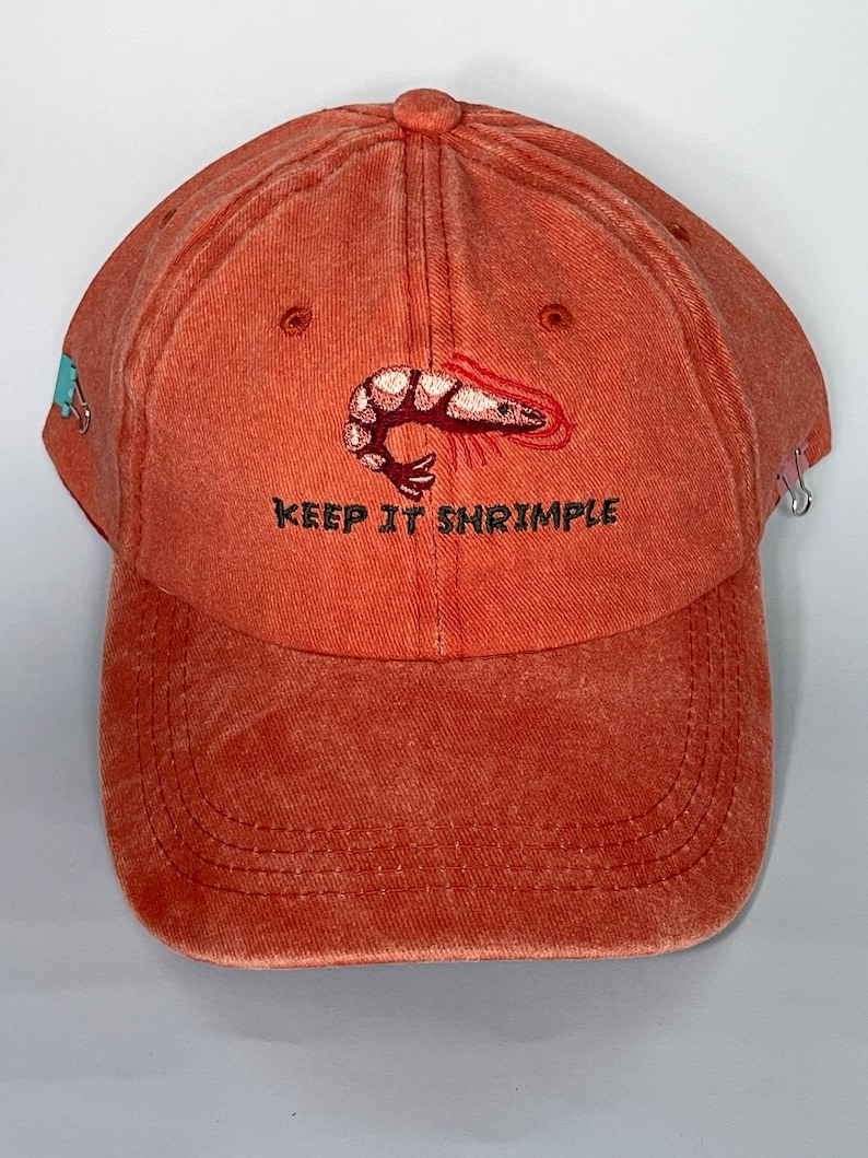 May include: A burnt orange baseball cap with a curved brim. The cap features an embroidered shrimp design and the phrase "KEEP IT SHRIMPLE" in black lettering. A small blue design is on the side.