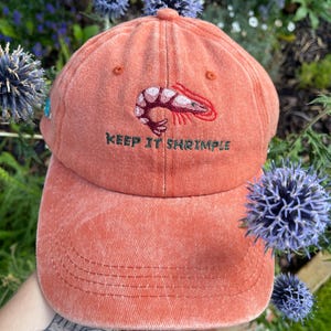 May include: A coral-colored baseball cap with a shrimp graphic and the text "KEEP IT SHRIMPLE" embroidered on the front. The cap has a curved brim and a classic six-panel design. The cap is made of a soft, textured fabric.