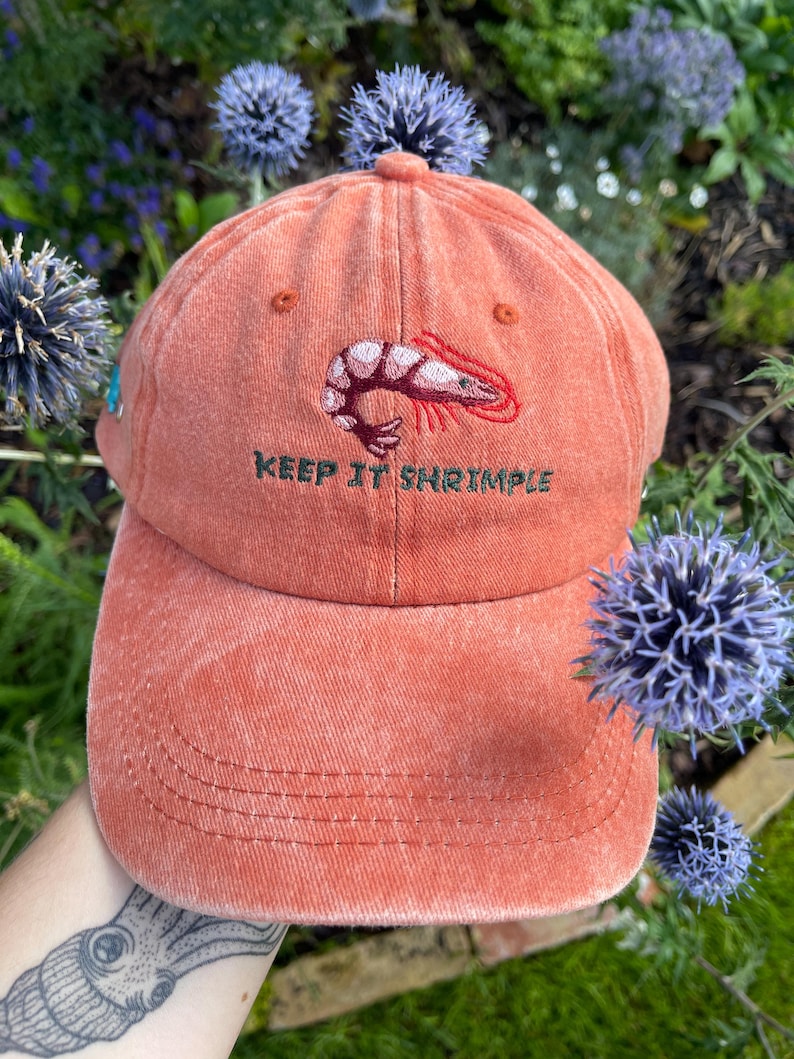 May include: A coral-colored baseball cap with a shrimp graphic and the text "KEEP IT SHRIMPLE" embroidered on the front. The cap has a curved brim and a classic six-panel design. The cap is made of a soft, textured fabric.