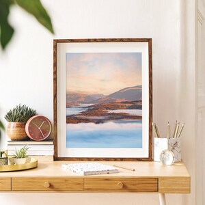 May include: A framed photograph of a lake with mountains in the background. The lake is covered in mist and the sky is a soft pink and orange. The frame is brown wood.