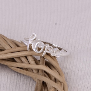 Puede incluir: Un anillo de plata con la palabra "hope" en cursiva. El anillo se muestra sobre un fondo neutro y un objeto circular tejido.