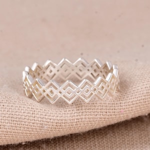 May include: A silver ring with a geometric diamond pattern. The ring is made of a shiny metal and sits on a beige fabric surface. The design features repeating open diamond shapes around the band.
