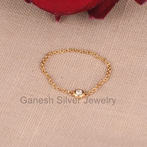 May include: A delicate gold chain ring with a single, clear round gemstone in the center.  The ring is on a beige fabric background. Ganesh Silver Jewelry.