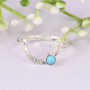 May include: A silver ring with a turquoise stone. The ring has a textured band and a curved design. The turquoise stone is round and set in a bezel setting. The ring is displayed on a white surface with white flowers in the background.