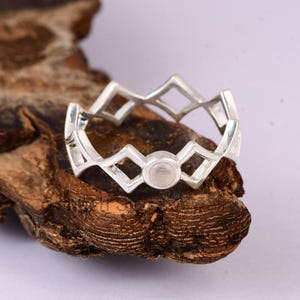 May include: A silver ring with a geometric design featuring diamond shapes and a central, round, light-colored stone. The ring is set against a textured, brown wooden surface, creating a contrast of textures and colors.