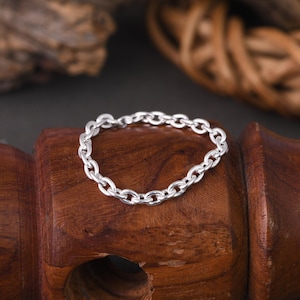 Puede incluir: Una pulsera de cadena plateada con eslabones ovalados, exhibida sobre una superficie de madera oscura. La pulsera está hecha de metal brillante y tiene un diseño simple y elegante. El fondo incluye elementos de madera y tejidos.