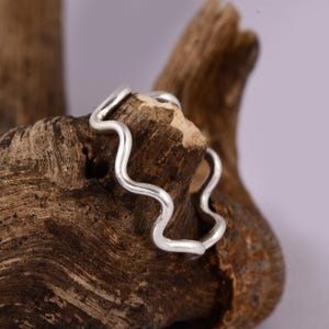 Può includere: Un anello in argento caratterizzato da un design ondulato o serpentino continuo. La fascia presenta una finitura metallica liscia e lucida e segue un motivo ondulato costante lungo tutta la circonferenza, creando una forma ritmica.