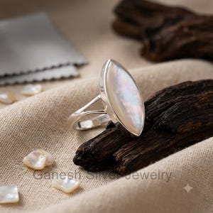 Könnte beinhalten: Ein Ring aus Sterlingsilber mit einem marquise-geschliffenen, irisierenden Perlmutt-Edelstein. Die Ringschiene ist mit 925 gestempelt, was auf Sterlingsilber hinweist. Der Ring hat eine geteilte Ringschiene und eine glatte, polierte Metalloberfläche.