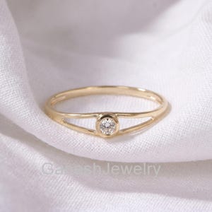 Op de afbeelding: Een gouden ring met een enkele ronde, briljant geslepen witte diamant in een zetting. De ringband splitst zich in twee dunne, parallelle armen die samenkomen bij de centrale steen, wat zorgt voor een open, geometrisch ontwerp met glans.
