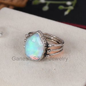 May include: A silver ring with a large, iridescent opal gemstone set in a textured, geometric bezel. The ring band is made of three thin bands with small silver beads on the sides.