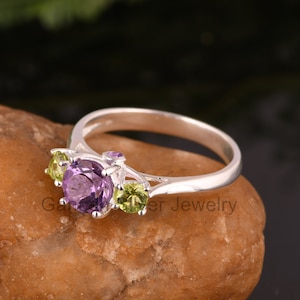 May include: A silver ring featuring a purple amethyst gemstone and two green peridot gemstones.