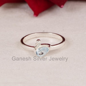 Może przedstawiać: Srebrny pierścionek z jasnoniebieskim kamieniem szlachetnym w kształcie łzy. Pierścionek leży na białej powierzchni z szarym napisem "Ganesh Silver Jewelry".