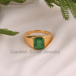 May include: A gold ring with a green gemstone. The ring has a square-shaped setting and a textured band. Ganesh Silver Jewelry is printed below the ring.