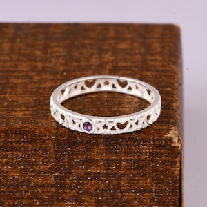 Könnte beinhalten: Ein silberner Ring mit einem filigranen Design mit Herzen und Sternen. Ein kleiner, runder, violetter Edelstein ist in der Mitte eingesetzt. Der Ring wird auf einer dunkelbraunen Holzoberfläche präsentiert.
