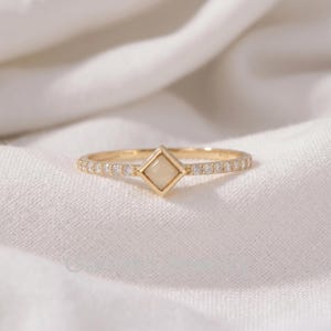 Puede incluir: Un anillo de oro con un engaste central en forma de diamante con una piedra opaca de color crema. La banda está decorada con una serie de pequeñas piedras transparentes de corte redondo incrustadas a ambos lados del punto focal cuadrado.