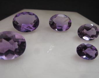 Könnte beinhalten: Fünf oval geschliffene Amethyst-Edelsteine in verschiedenen Größen, jeder mit einer facettierten Oberfläche, die das Licht reflektiert. Die Steine sind tiefviolett und in einem lockeren Kreis angeordnet, was ihre Transparenz unterstreicht.